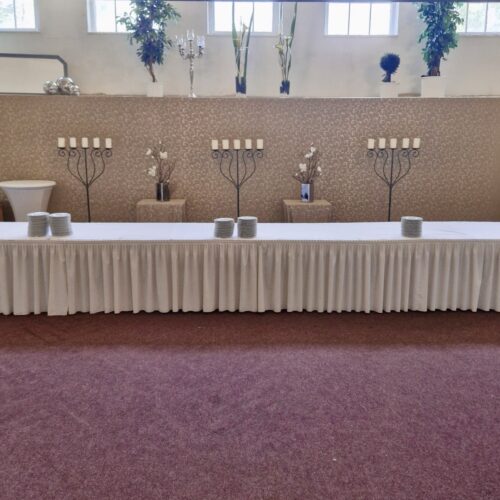Langer, mit weißem Tuch bedeckter Tisch vor dekorativer Wand mit Kerzenhaltern und Blumenarrangement.